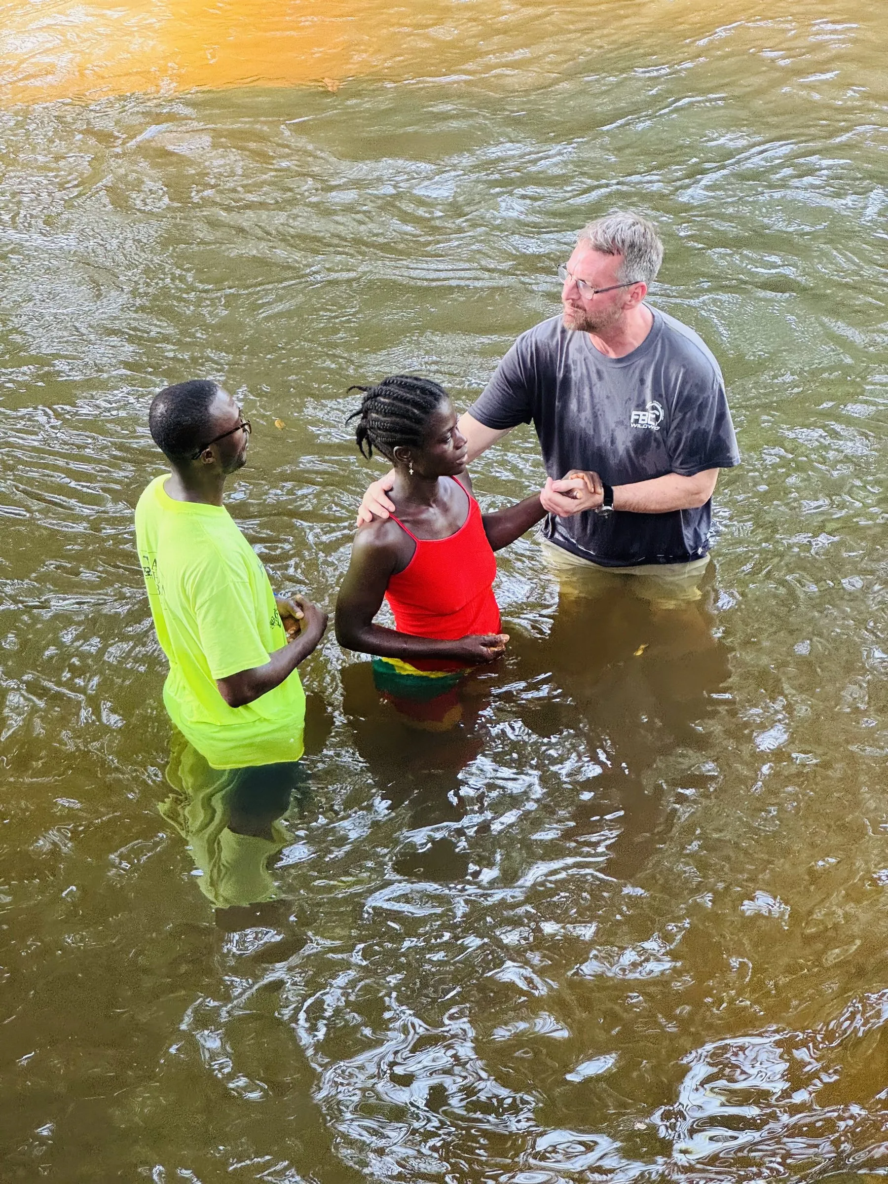 A young woman being baptized in a river, supported by two men standing waist-deep in the water beside her.