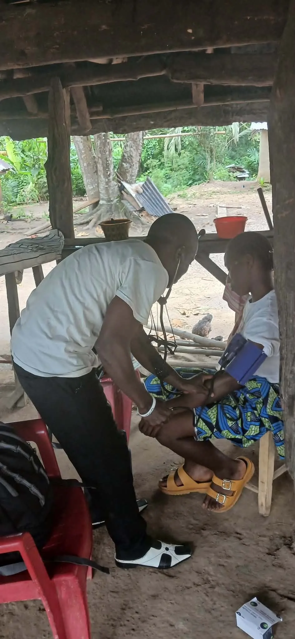 A HOPE127 health worker taking a young woman's blood pressure, seated under an open shelter in a village.
