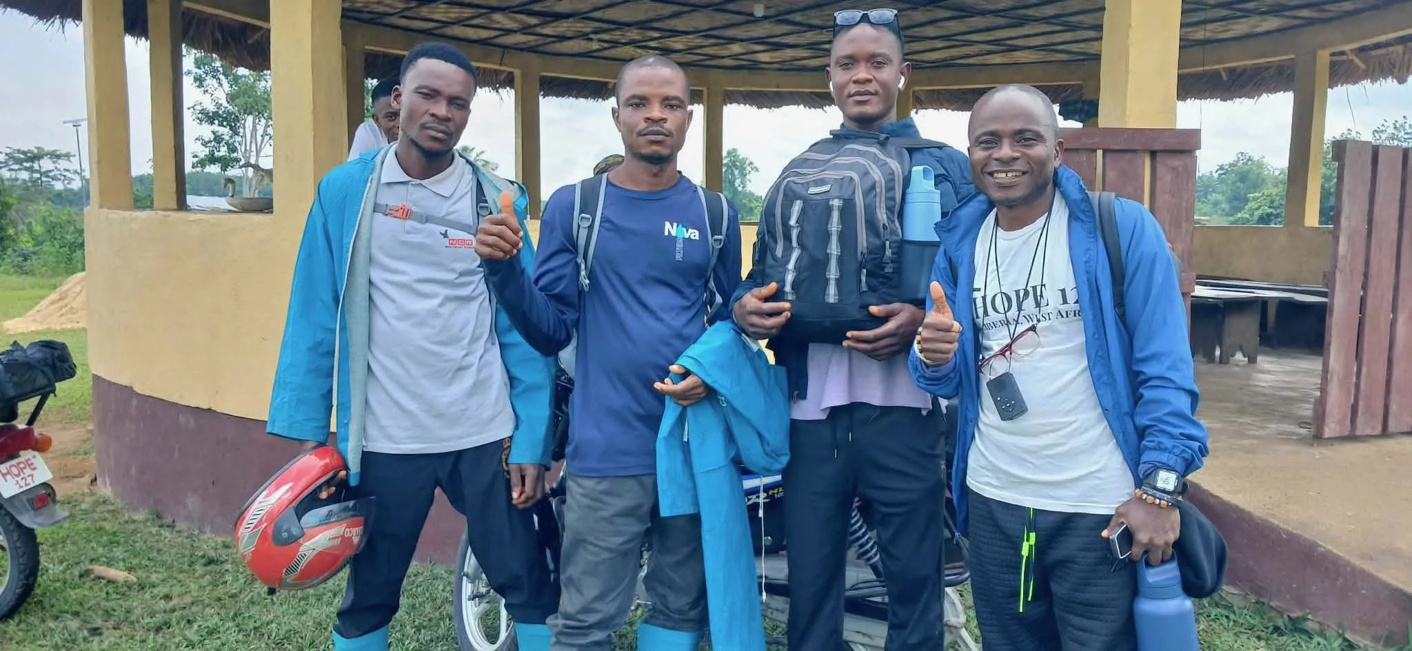 Four Liberian missionaries standing together at an open-air village shelter. One wears a HOPE 127 Liberia West Africa t-shirt; a motorbike and a bicycle helmet are beside them.
