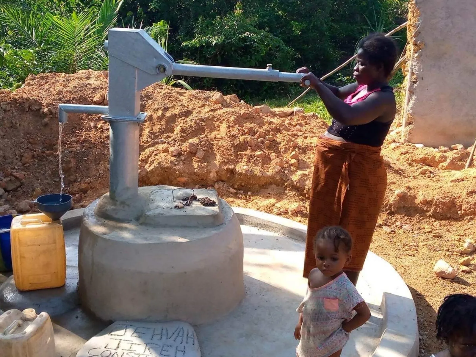 A woman working the handle of a hand-pump well, water running from the spout into a basin, a small child standing beside her on the well's concrete apron and jerrycans waiting to be filled.