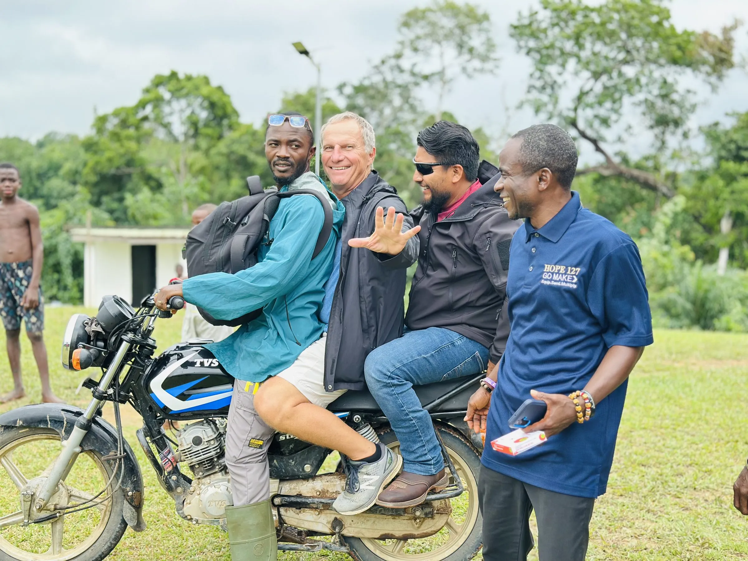 Four men gathered around a motorbike on grass, two of them seated on it, one wearing a navy HOPE 127 polo shirt reading GO MAKE — Equip. Send. Multiply.