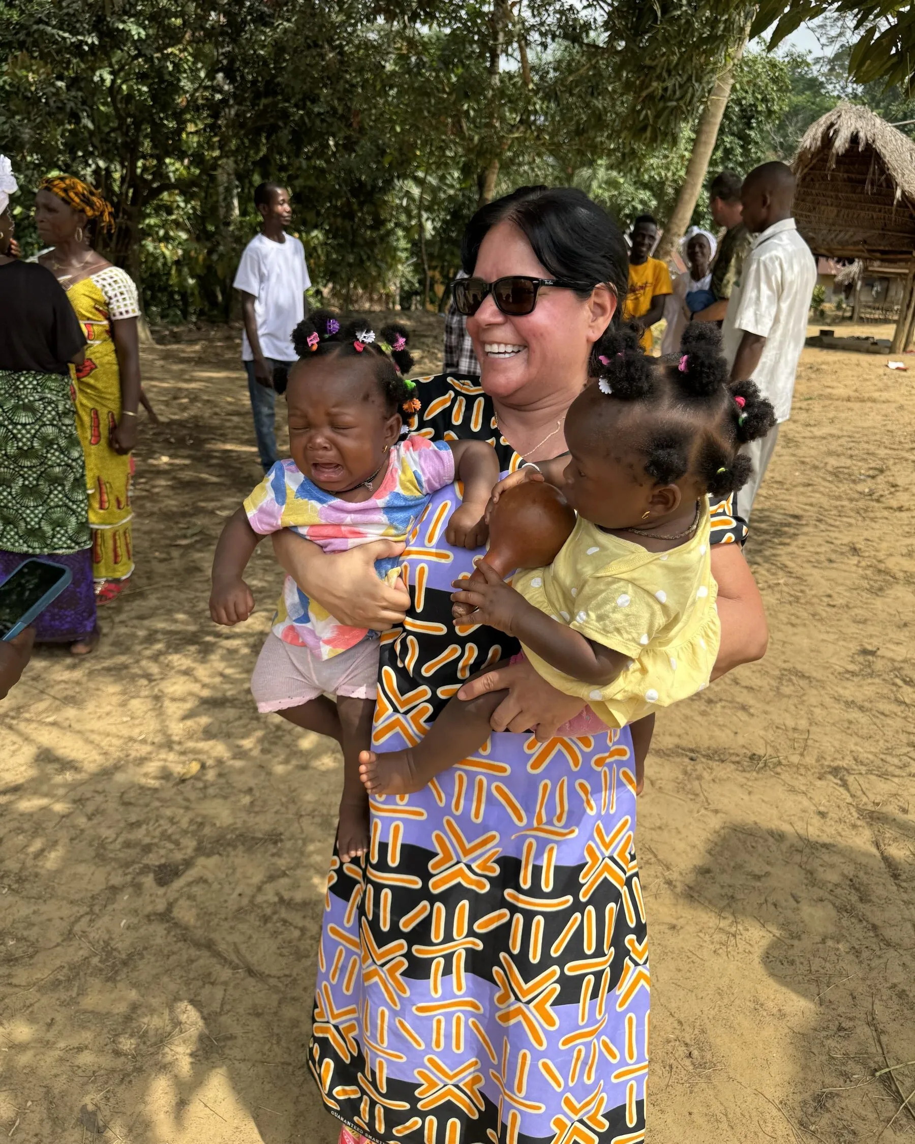 Anamaria Bohorquez standing in a village clearing holding two toddlers, one on each arm, with villagers and a thatched roof behind her.