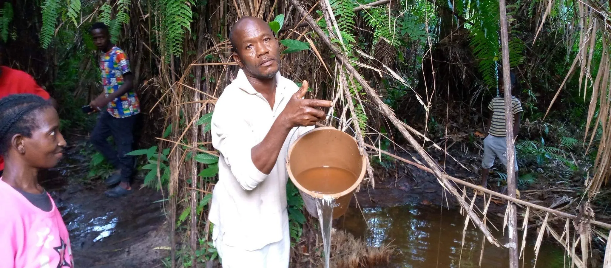 A man standing in a creek bed, holding out a bucket and pouring brown water so that it catches the light.