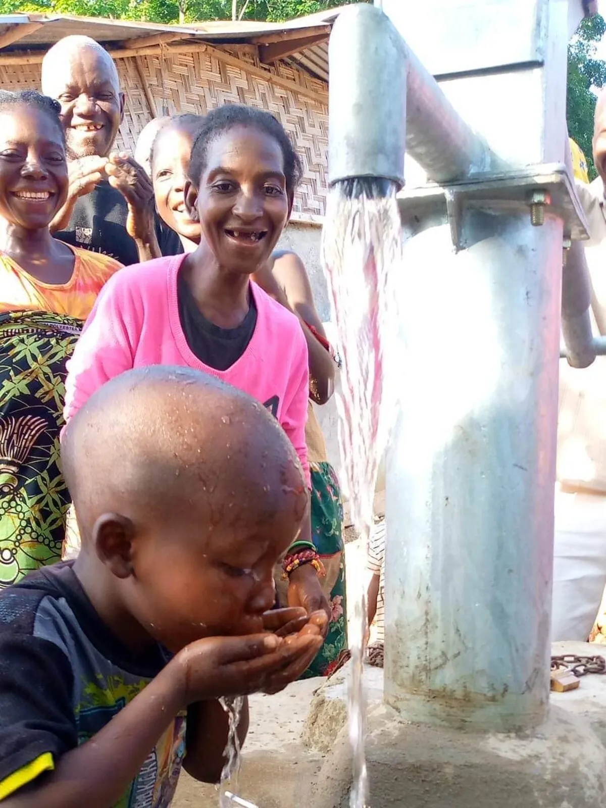 A small boy drinking from his cupped hands under a running hand pump, his family standing behind him and laughing.