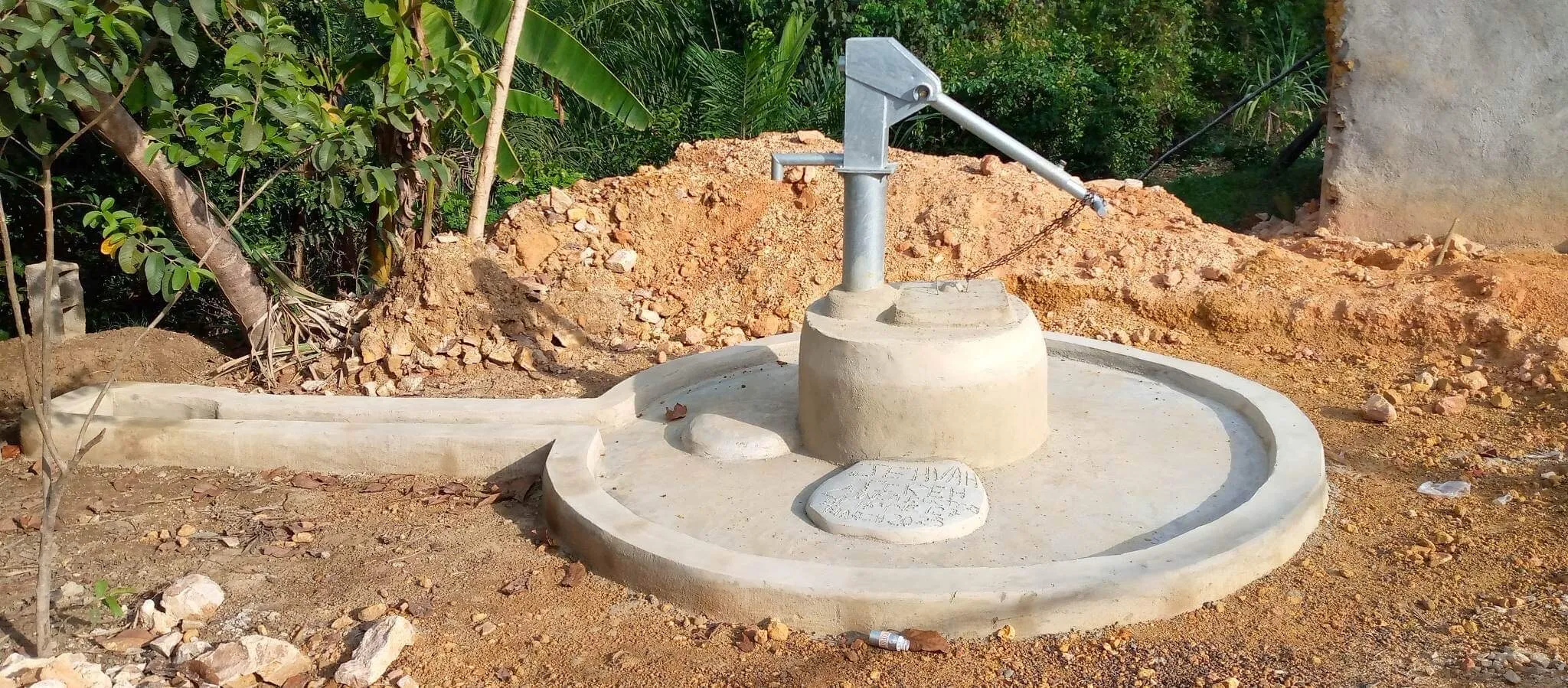 A hand-pump well standing on a fresh concrete apron with an inscribed plate set into it, red earth banked up behind.