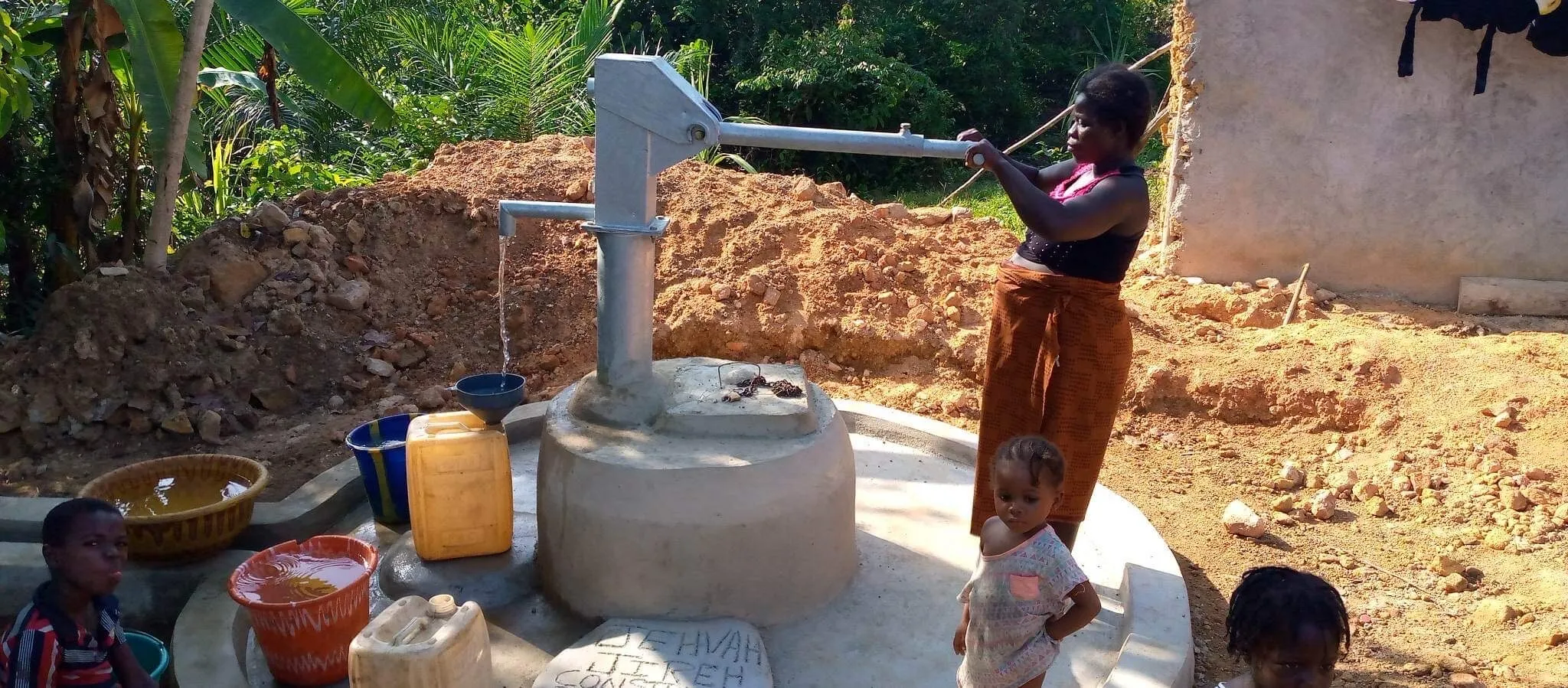 A woman working a hand pump with a small child beside her, jerrycans and basins filling on the concrete apron.