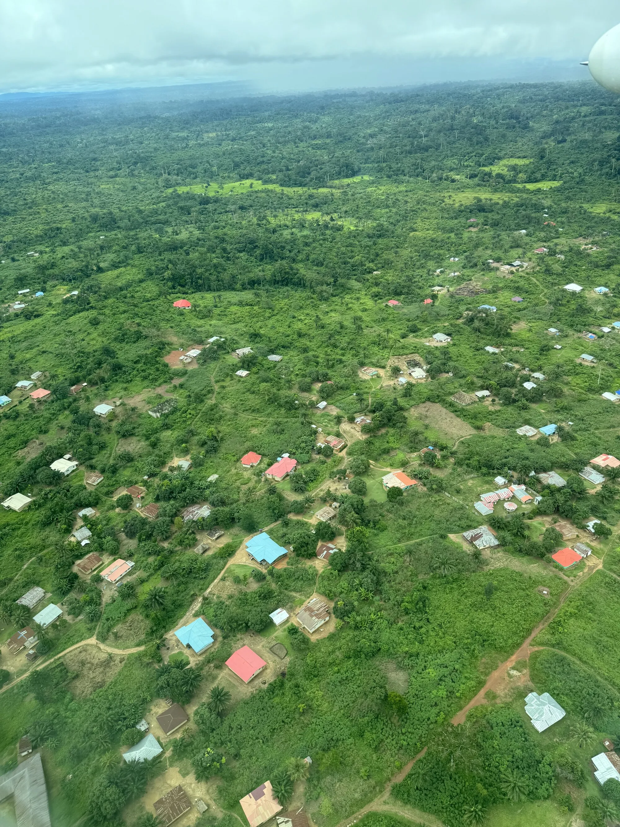An aerial photograph of Liberia taken from a plane window: dense green forest running to a hazy horizon under low cloud, and below it a village of houses roofed in red, blue and bare metal, scattered among clearings and joined by red-earth footpaths.